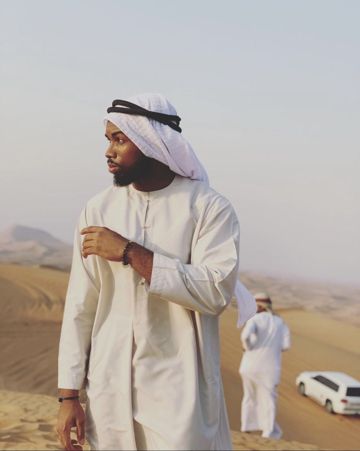 OJ in a white kandura on a sand dune, a 4x4 parked below